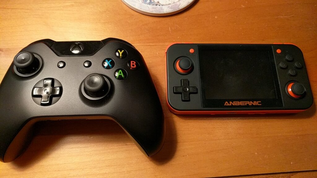 An Xbox One controller, laying on a table next to an Anbernic RG-350 handheld.  The two devices are similar in size.