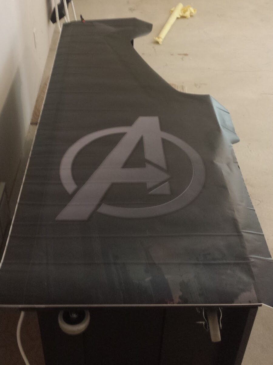 A sheet of black decorative vinyl adhesive with an Avengers "A" logo is laid loosely on the side of the arcade cabinet, ready for installation 