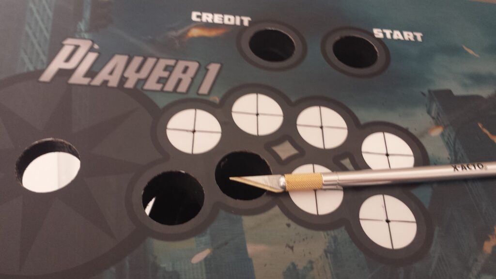 An X-acto Knife lays on top of the installed control panel artwork.  Five of the 11 visible button & joystick holes have been cut away from the art, so the controls can be installed.