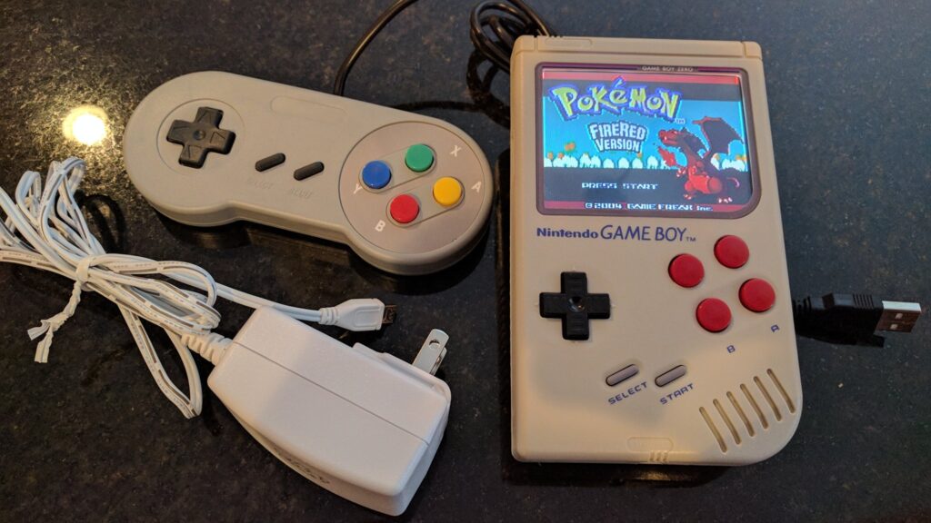 A Game Boy Zero handheld, with Pokemon FireRed displayed on the screen.  Also pictured are a USB SNES-style controller, and an AC adapter/charger.