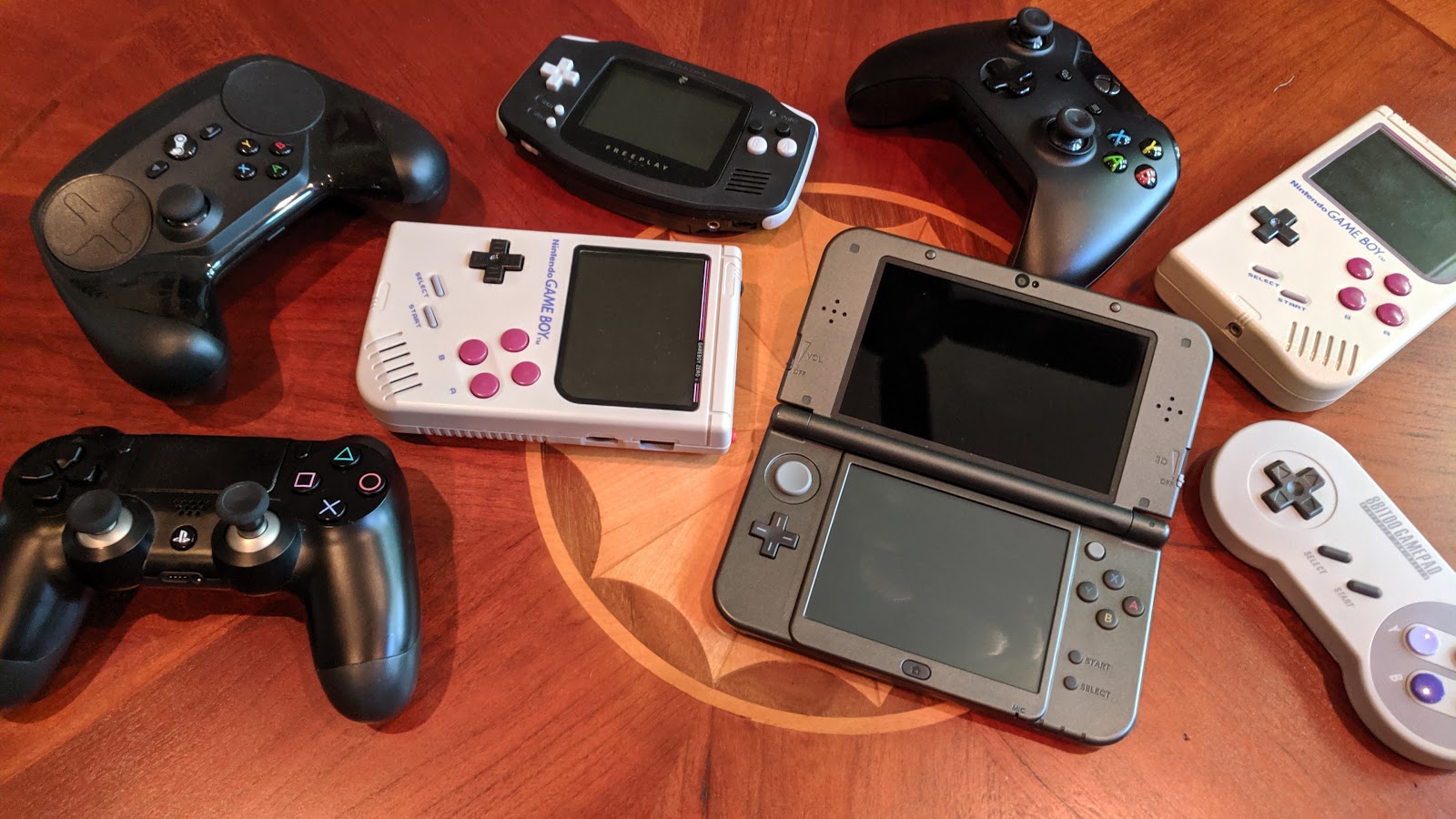 A collection of handheld game consoles and controllers. Included are a Steam Controller, a DualShock 4, two classic Game Boy-based handhelds, a classic Game Boy Advance-based Freeplay Advance, an Xbox One controller, a New Nintendo 3DS XL, an an 8BitDo SNES30 controller.
