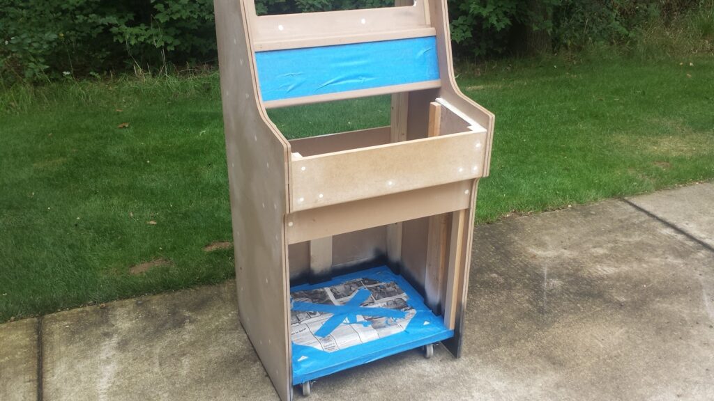 Front view of a partially assembled arcade cabinet.  Screw holes have been filled & sanded, and painters tape covers the administration panel & inside bottom surface.