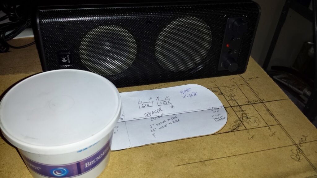 A PC speaker sitting on top of a section of MDF. On the MDF are markings, laying out where to cut an opening for audio to pass through the speaker panel.